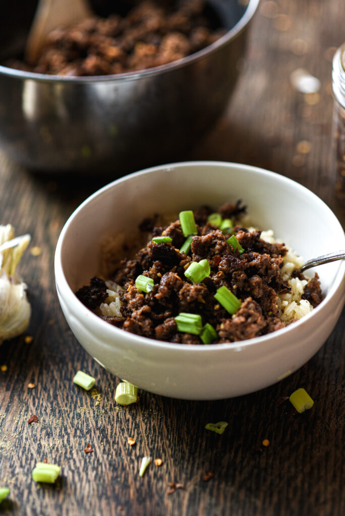 A bowl of Korean beef over rice, topped with chopped green onions, sits on a rustic wooden surface. In the background, a metal bowl of cooked ground beef and a jar of chili flakes or sauce are visible. A garlic bulb rests nearby. Text overlay at the bottom reads: “From My Kitchen: Korean Beef Bowls.”